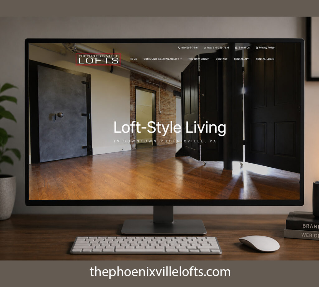 Wide computer monitor showcasing The Phoenixville Lofts apartment website homepage in a contemporary office setting with warm lighting and modern desk accessories.
